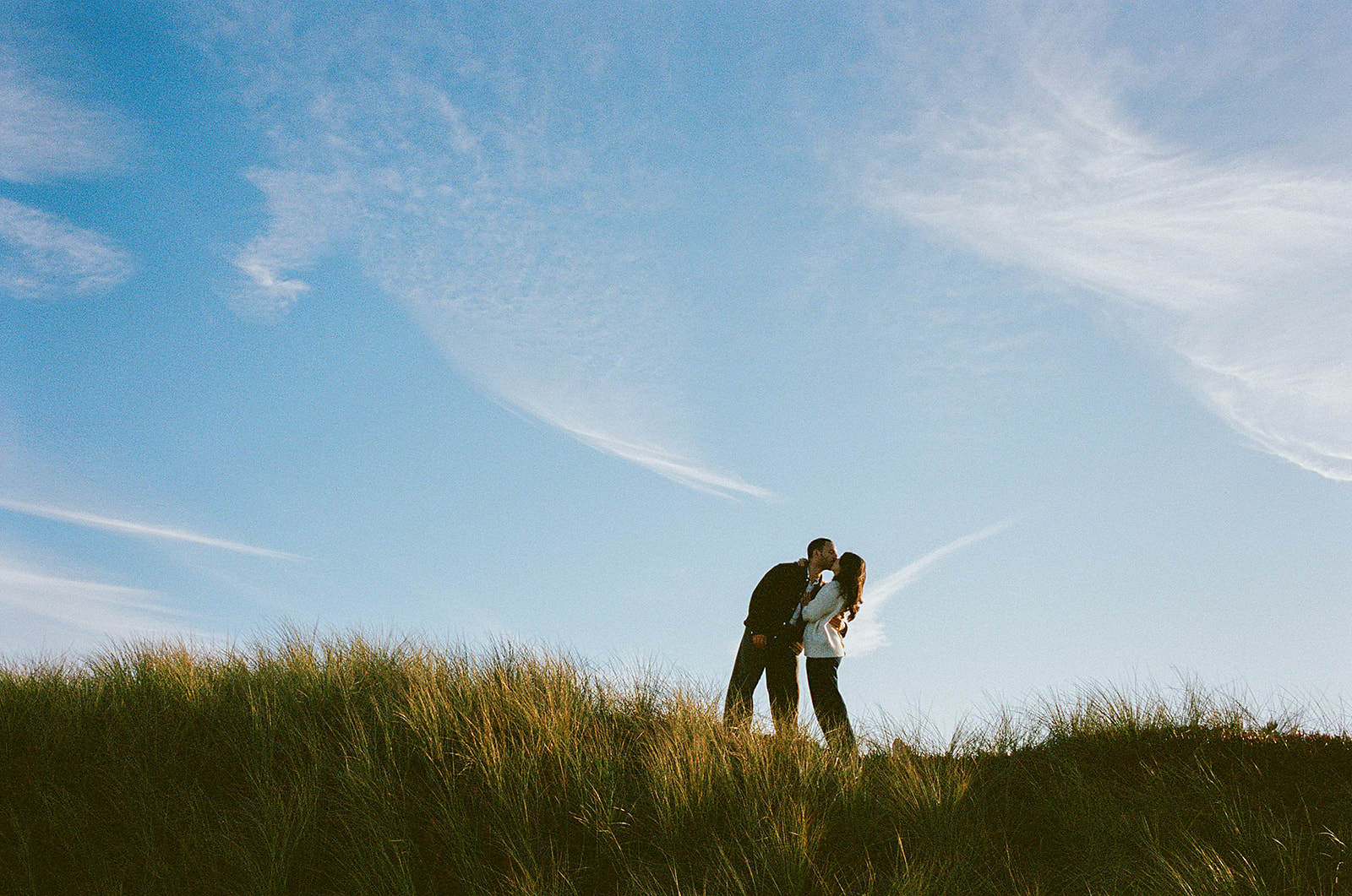 Couple kissing on grassy sand dunes during a Point Reyes engagement session photographed on film along the Northern California coast.